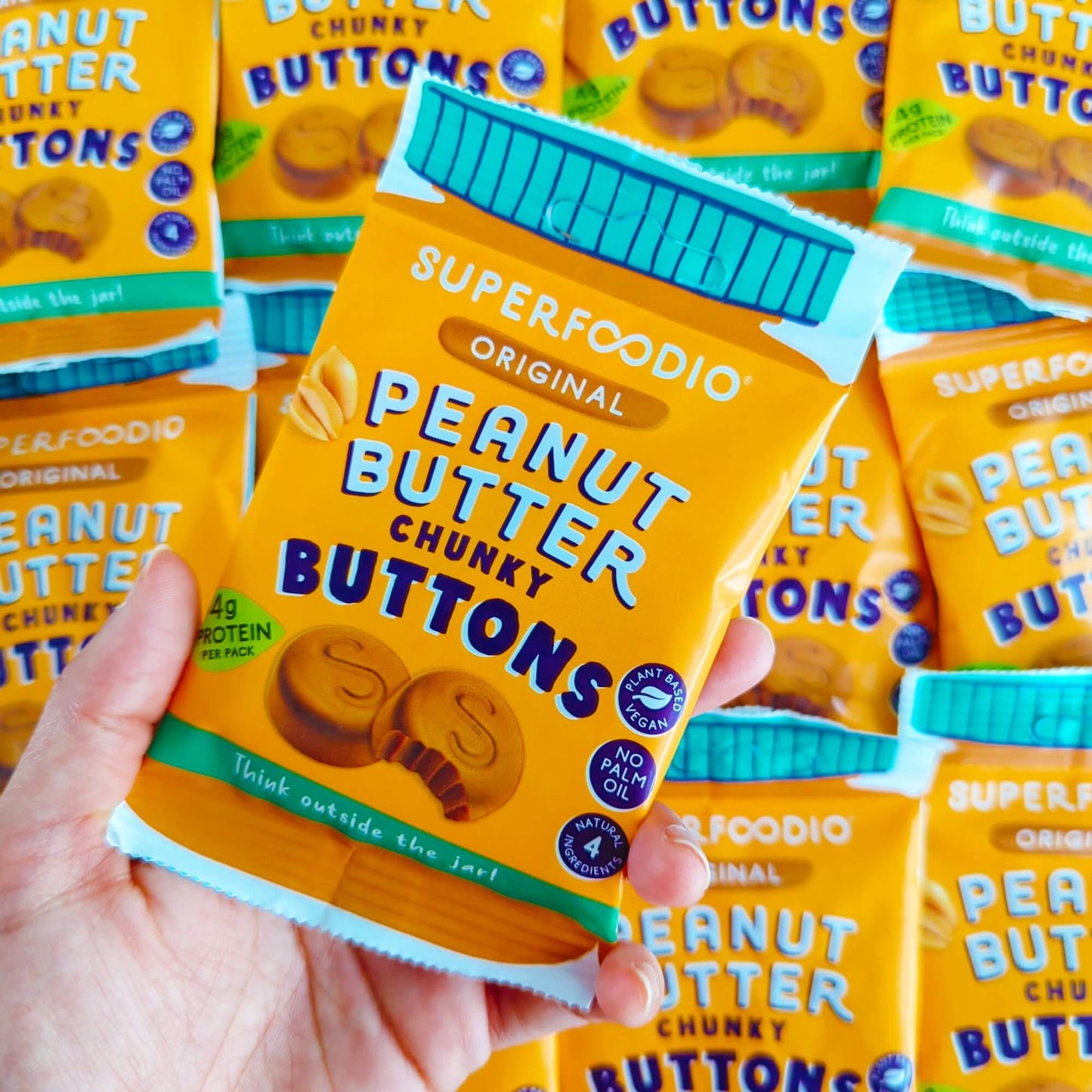 Original Peanut Butter Butons 15 x 20g - Superfoodio. Hand holding a single bag over the top of a pile of bags
