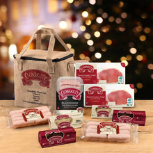 A Clonakilty food gift set arranged on a wooden surface in front of a blurred Christmas tree with warm bokeh lights. The set includes a beige insulated tote bag, multiple packages of Clonakilty sausages, bacon, black pudding and white pudding, all branded with the Clonakilty logo.