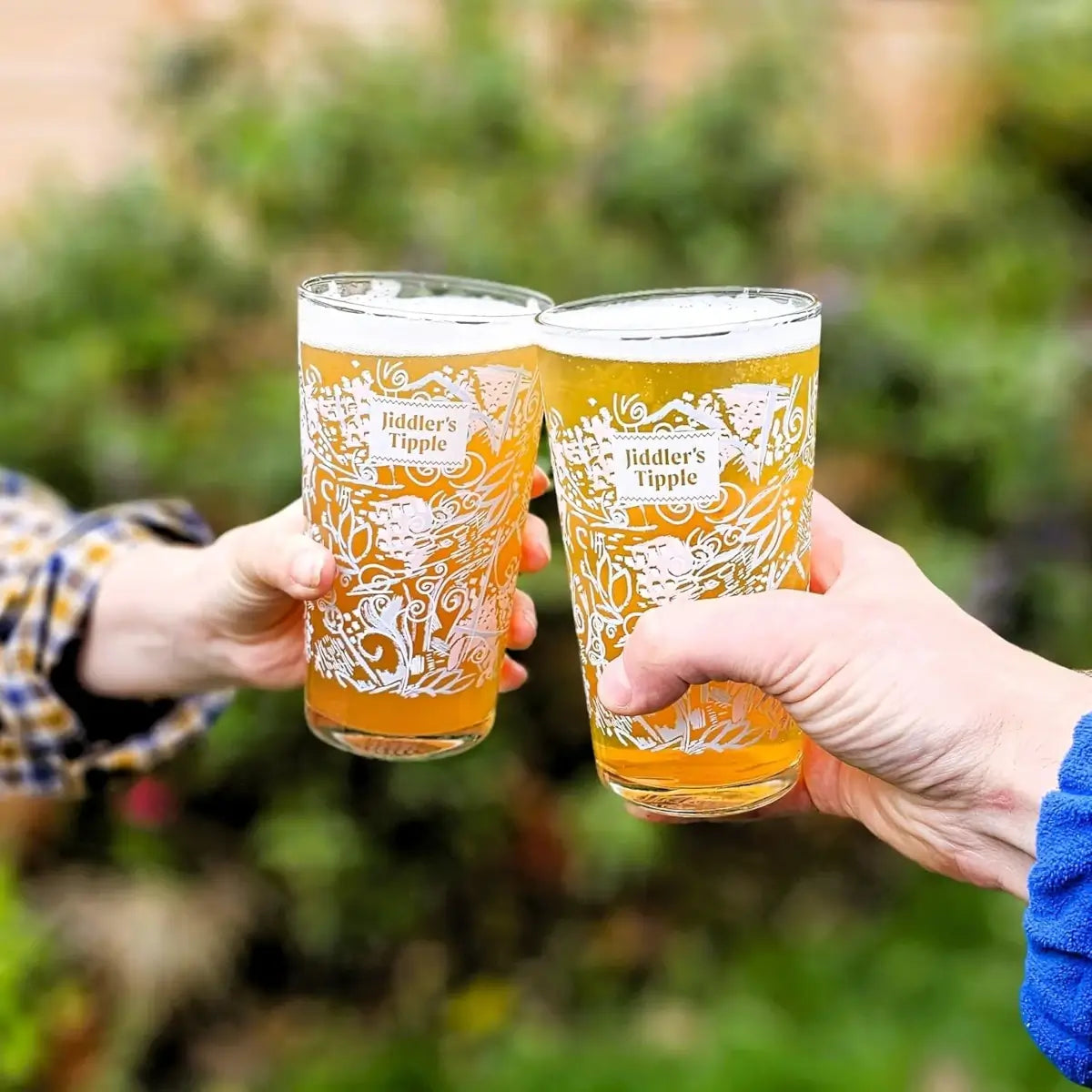 Two hands holding Jiddler's Tipple glasses outdoors with greenery in the background