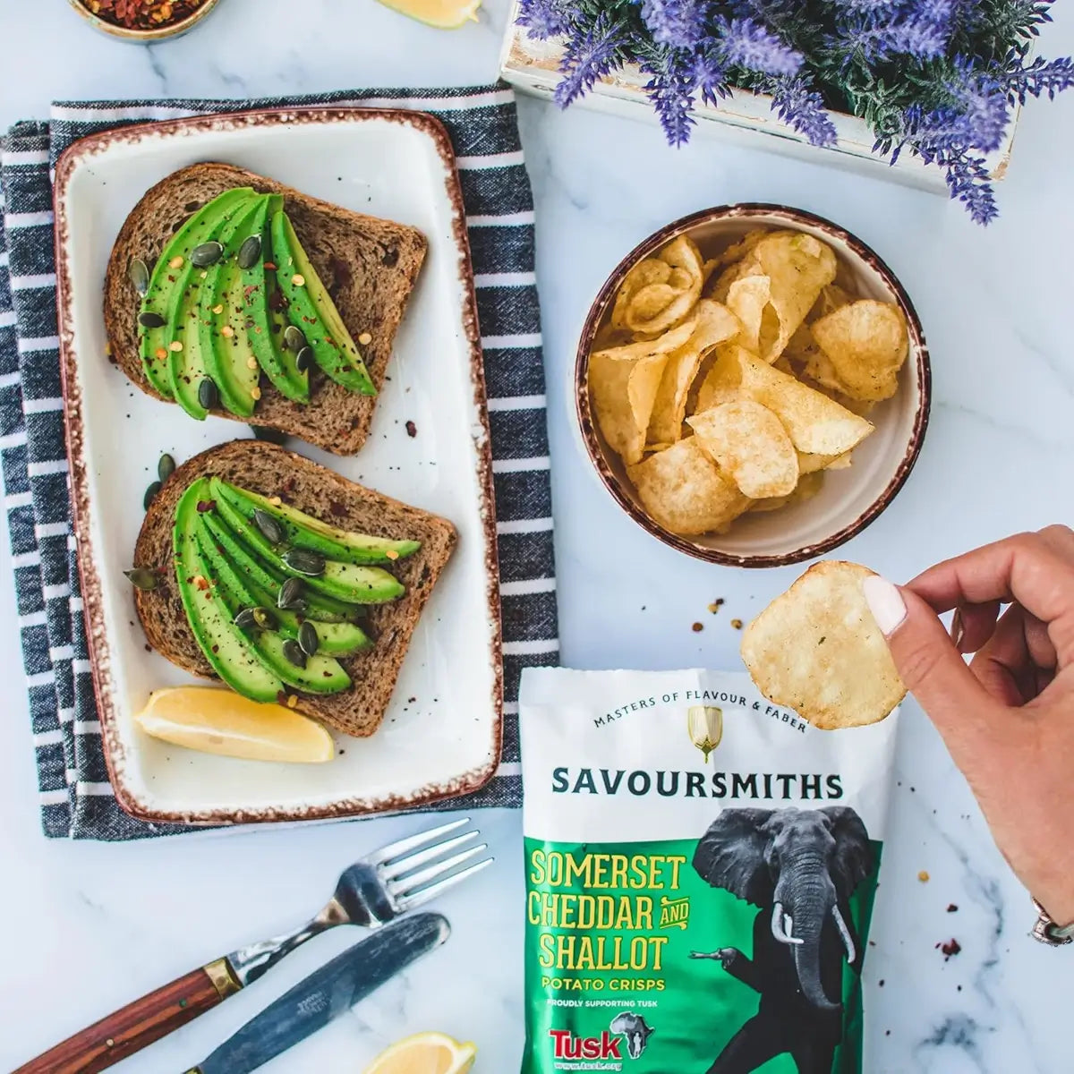 Savoursmiths  - Somerset Cheddar and Shallot Luxury Potato Crisps next to a plate of with 2 brown toasts, topped with avocado and lemon
