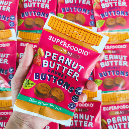 Peanut Butter & Jam Buttons 15 x 20g - Superfoodio. Hand holding a bag of the buttons above a pile of bags.